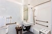 Accessible guest bathroom with single lighted vanity area, toilet and shower with grab bars, and portable chair.