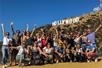 A large group of hikers wearing sunglasses posing for a group photo with the Hollywood sign in the background.