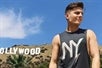 A man wearing a gray NY tank top looking to the right on a sunny day with the Hollywood sign in the background.