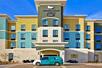 The blue, yellow, and gray front exterior of the Homewood Suites by Hilton New Braunfels with a blue van parked out front on a sunny day.