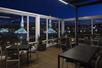 A rooftop patio with glass walls and no roof, several black square tables with matching chairs, and a great view of the city of Portland at night.