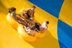 Four young adults in a yellow tube going down a yellow and blue checkered waterslide on a sunny day.