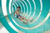 A young boy in blue swim trunks sliding down an aqua and clear striped water slide while smiling.