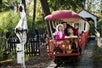 A father and his two daughters in costumes riding at the front of a red train ride with headstones and other spooky decorations to the side of them.