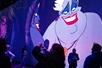 People in a darker room with illuminated walls with a large illustration of Ursula from "The Little Mermaid," with a sinister smile and dramatic makeup.