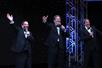 Three men in tuxes smiling and holding microphones to their faces while waving with their other hand on stage at the Jew Man Group Comedy Show.