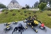 A colorful Lego scene at Legowood, featuring a variety of large Lego spiders on a grassy hill, with a whimsical building in the background and the "LEGOWOOD" sign prominently displayed.