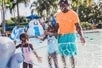 A joyful scene of a man and two children playing in a water park, splashing water and enjoying a sunny day amidst colorful slides and palm trees.