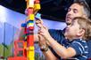 A father and son smiling and building a tower out of colorful LEGO at LEGO Discovery Center Boston.