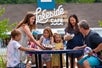 A family enjoying a meal together at an outdoor cafe named "Lakeside Cafe," with the adults sharing food from a paper container with the children.