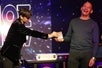 Lioz in all black performing a trick on stage with a bald man in a long sleeve shirt who is laughing at his show in Las Vegas.