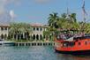 A red & black Pirate Ship in blue water cruising in front of a stunning mansion with palm trees surrounding it on a sunny day in Miami.