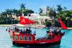 A bright red pirate ship with two upper desks full of passengers on a sunny day with a large white house and palm trees in the background.