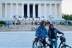 Two women in jackets posing for a photo with their bikes in front of the Lincoln Memorial with tons of tourists behind them on the steps.