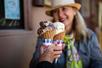 A woman wearing a hat and floral scarf holding her waffle cone up to someone another persons and smiling.