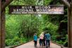 A group of people walking of a wooden bridge leading into the woods with the wooden arch sign for the Muir Woods National Monument behind them.