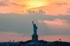 The Statue of Liberty with puffy clouds and a pink and orange sunset behind it on the NYC Skyline Sunset Cruise.