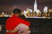 View of a man and woman hugging from behind while near the railing of a cruise boat on the New Jersey Dinner Cruise with the city in the distance.