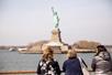 Three women leaning on the railing of a cruise boat while looking at the Statue of Liberty in the distance on a sunny day.