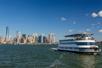 A large white cruise boat in the water with New York City in the back ground on a sunny day with a bright blue sky overhead.
