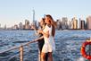 Two young women stranding near at the railing on the top deck of a cruise boat with New York City in the background and the sun shining on them.,