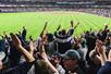 Yankees fans in the stands cheering and jumping with their arms in the air with the bright green field in front of them.