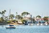 A lone sail boat sitting in the water with a marina full of boats behind it on a sunny day in Newport Beach, California.