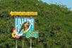 A teal, white, and yellow Haleiwa North Shore signage with the sun shining on it and trees in the background.