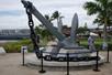 The USS Arizona Memorial Anchor  on display with a rope guard around it and people walking by in the background on a sunny day.