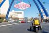 View of the back of a yellow GoCar driving down the road with a pink Las Vegas sign hanging over the road on a sunny day.