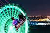 A young man and woman on a swing ride in motion at night with a green glowing ferris wheel in front of them.