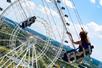 A woman sitting in a swing ride seat by herself in the air with an empty seat and a ferris wheel in front of her of a sunny day.