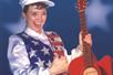 Patty Waszak in an American Flag vest and a white hat giving a thumbs up and holding a red guitar.