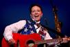 Patty Waszak in a white shirt and American Flag sequin vest sining and playing a red guitar.