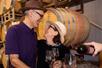 An older couple wearing fedoras looking at each other and smiling with wine being poured in front of them and barrels of wine behind them.