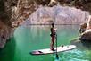 A woman standing on a paddle board at the mouth of a cave with green water under her and the sun shining in the background.