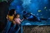 A woman and a young girl sitting in front of an aquarium, joyfully interacting with a diver underwater who is reaching out to them, surrounded by colorful fish and a sea turtle.
