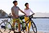 A man and woman wearing helmets and walking their bicycles on a beach with the sun shining on them and water in the background.