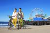 A man and woman wearing helmets posing with the bicycles in the sand near the ocean with Santa Monica Ferris Wheel in the background.