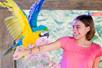 A young girl in a pink shirt smiling with her arm out and a blue and yellow macaw landing on it at SeaQuest.