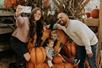 A mother and father taking a selfie with their two young children who are sitting in a pumpkin chair with dried corn stalks behind them.