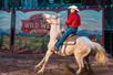 A man in a red shirt and white hat riding a white speckled horse and pulling on the reigs.