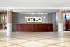 A long dark wooden front desk with three light over it and art on the wall behind it in a lobby with beige stone floor.