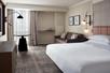 A hotel room with a white king bed, brown sofa, table and chairs, dresser, TV, and a small window.