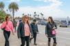 A group of women wearing black and pink walking on the sidewalk with a blue sky over them and lots of palm trees behind them in San Diego.