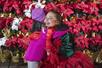 A Six Flags cast member in a green top and red skirt hugging a young girl in a bright pink coat with a wall of poinsettias behind them.