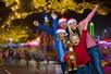A family of four wearing Santa hats posing for a photo at Six Flags during Holiday in the Park at night with Christmas lights behind them.