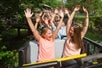 A group of adults and kids revel in the thrill of the ride, their arms raised in excitement, as they board the River King Mine Train on an early afternoon, with lush trees and a wooden fence in the background at Six Flags St. Louis.