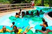 A lazy river full of people floating on clear green tubes while laughing and having a good time on a sunny day.