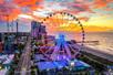 Aerial wide shot of the Myrtle Beach SkyWheel with a bright orange sunset behind it and the lively city to the left.